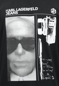 Μαύρο πουκάμισο με πορτραίτο σε λευκό ημιτόνο που φορά γυαλιά ηλίου, vintage κάμερα, σκίτσο με στυλό και το κείμενο "KARL LAGERFELD JEANS".
