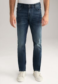 Dunkelblaue Denim-Jeans mit schmaler Passform, klassischem Fünf-Taschen-Design und leichter Ausbleichung an den Oberschenkeln. Kombiniert mit weißen Sneakers.