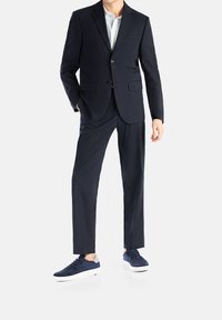 Marineblaues Anzug mit passender Hose, taillierte Passform, Blazer mit zwei Knöpfen und einer Tasche; kombiniert mit marineblauen, strukturierten Sneakers und weißen Sohlen.