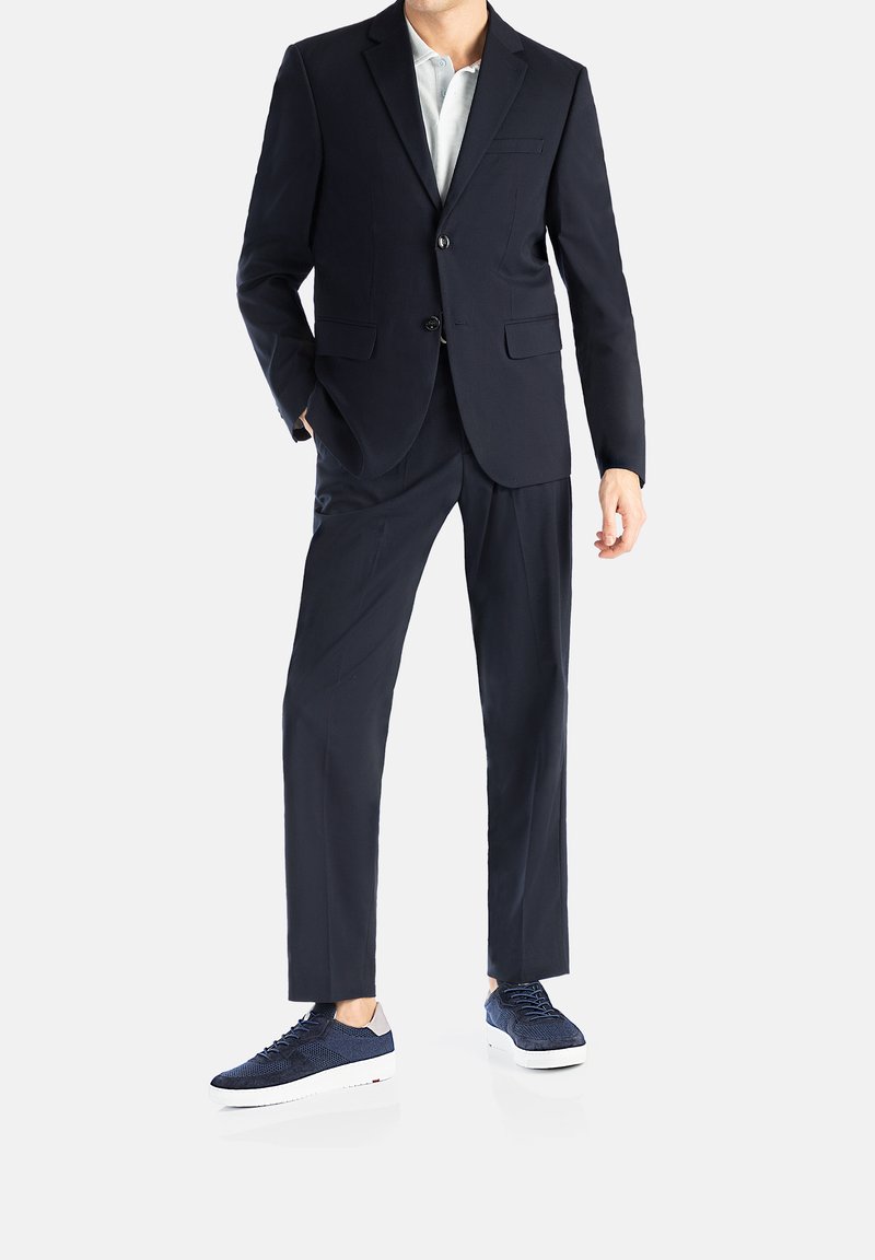 Marineblaues Anzug mit passender Hose, taillierte Passform, Blazer mit zwei Knöpfen und einer Tasche; kombiniert mit marineblauen, strukturierten Sneakers und weißen Sohlen.