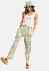 Pantaloni floreali verde chiaro con motivi tropicali rosa e gialli, abbinati a una canotta bianca e a un cappello a secchiello verde. Sandali a piattaforma color sabbia.