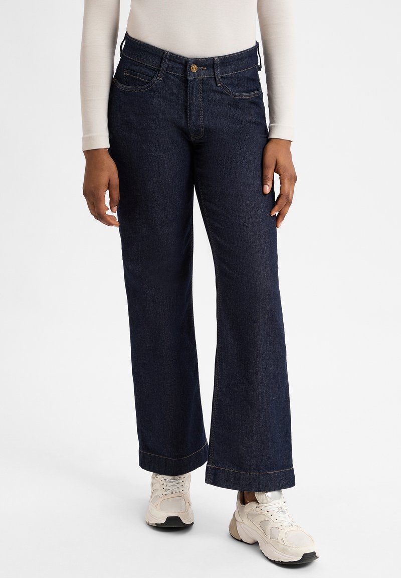 Jean en denim bleu foncé à jambe large et taille haute, avec poches frontales et fermeture à bouton doré. Associé à des baskets beiges.