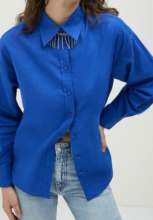 Personne portant une chemise boutonnée bleu vif avec un col décoratif orné de perles et un jean en denim bleu clair, main sur la hanche.