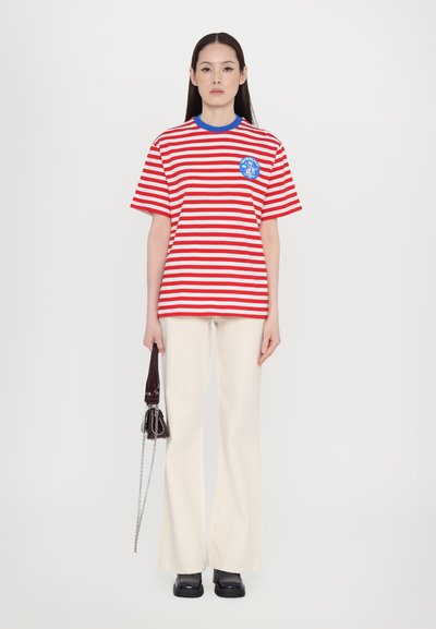 Red and white striped t-shirt with blue collar, featuring a round logo patch. Paired with cream flared pants and a small dark handbag.