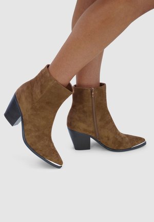 Bruine suède enkellaarsjes met een spitse neus, een zijrits en een lage blokhak. De zool heeft een zilveren accent bij de neus.