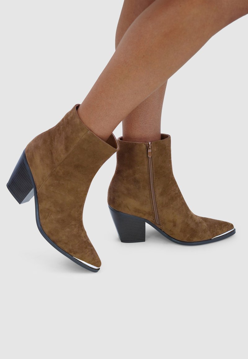 Bruine suède enkellaarsjes met een spitse neus, een zijrits en een lage blokhak. De zool heeft een zilveren accent bij de neus.