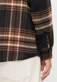 Brown and black plaid flannel shirt, featuring a soft texture, buttoned cuffs, and side slits, paired with cream-colored pants.
