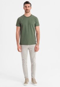Muž stojící a oblečený v zeleném vzorovaném tričku s krátkým rukávem, světle šedých slim-fit kalhotách, béžových teniskách a náramku z korálků na pravém zápěstí.