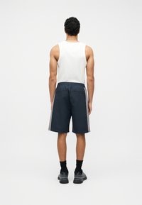 Άνδρας όρθιος, γυρισμένος πλάτη, φοράει λευκό αμάνικο μπλουζάκι, σκούρο μπλε σορτς μ ε γκρι ρίγες στα πλάγια, μαύρες κάλτσες και μαύρα αθλητικά παπούτσια σε απλό φόντο.