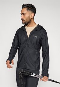 Fekete cipzáros dzseki struktúrált kapucnival, könnyű anyagból, oldalsó zsebekkel és hüvelykujjnak való réssel; túrabotokkal viselve.