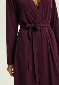 Głęboko burgundowa szata wykonana z miękkiego materiału, charakteryzująca się krojem typu wrap, długimi rękawami i wiązanym pasem. Widoczne detale koronkowe przy dekolcie.