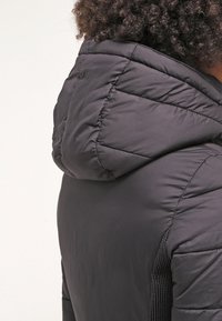 Svart vadderad jacka med hög krage, quiltade sömmar och texturerade sidopaneler. Mjuk, lätt och isolerad material.