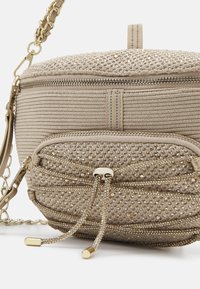 Beige strukturierte Umhängetasche mit goldener Kettenriemen, Reißverschlussverschluss und vorderem Fach, das mit kleinen glitzernden Perlen und Zugkordeln verziert ist.