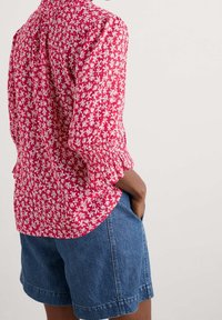 Rød blomsterbluse med trekvartærmer, der har en smocket manchet og en afrundet kant, parret med blå denimshorts.