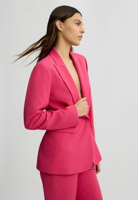 Blazer rose vif avec un col à revers, fermeture à un bouton et manches longues, associé à un pantalon assorti, présenté sur un mannequin.