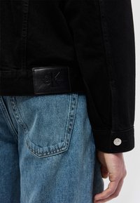 Κοντινό πλάνο ενός ατόμου που φοράει ανοιχτόχρωμα γαλάζια τζιν και μαύρο μπουφάν με μαύρο δερμάτινο λογότυπο Calvin Klein κοντά στη μέση.