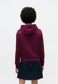 Felpa con cappuccio bordeaux con coulisse, polsini e bordo a coste. Abbinata a una gonna in denim scuro, che mostra un look casual a strati visto da dietro.