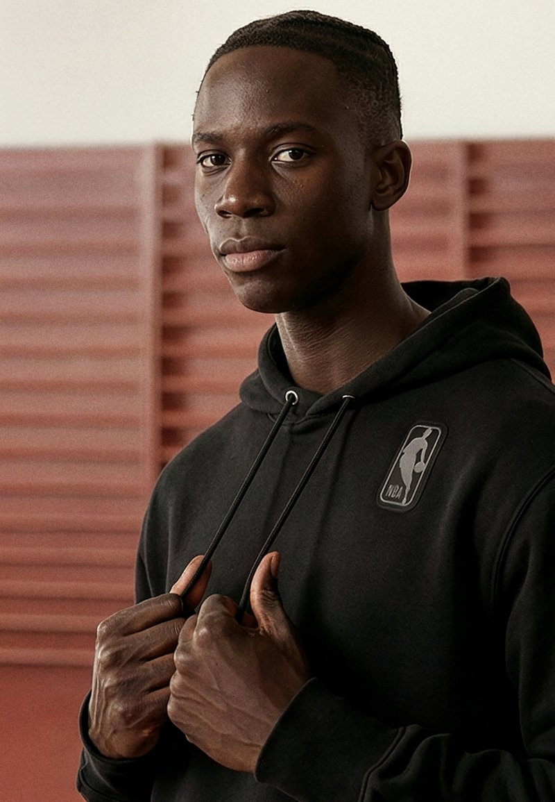 Jeune homme portant un sweat à capuche noir NBA, ajustant les cordons avec une expression neutre, debout devant un fond en bois.