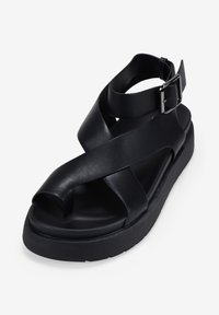 Sandalo in pelle nera con cinturini incrociati e fibbia alla caviglia. Suola spessa con una leggera curvatura, dotato di un design aperto sulla punta e una texture liscia.