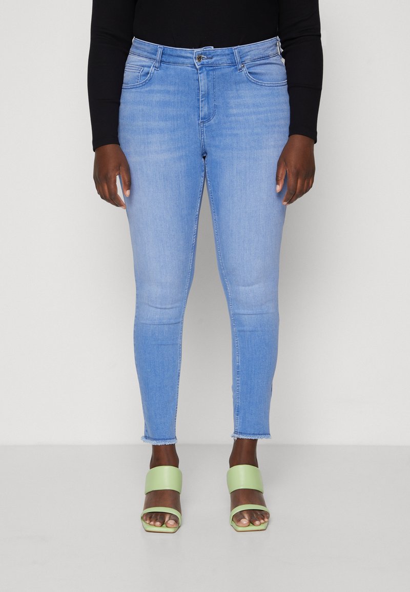 Light blue skinny jeans with a frayed hem, five-pocket design, and a smooth texture, paired with green block-heeled sandals.