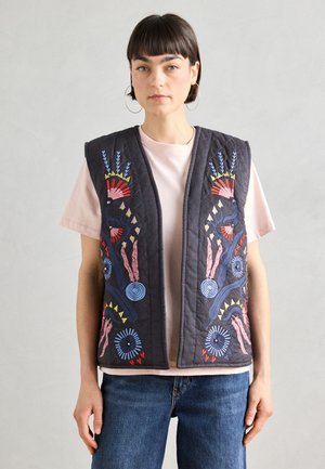 Gilet trapuntato nero con ricami colorati a motivo floreale e astratto, bottoni rotondi e una vestibilità comoda. Indossato sopra una maglietta rosa chiaro.