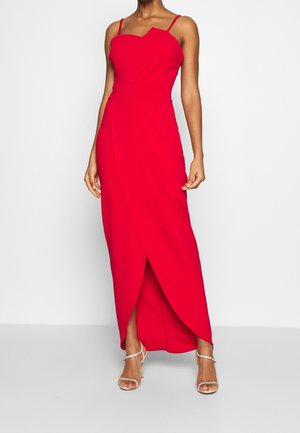 Femme portant une robe maxi rouge sans manches avec des bretelles fines et des sandales à talons hauts argentées, debout devant un fond blanc.