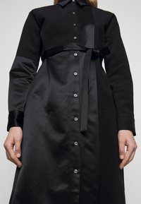 Svart klänning i slätt tyg med knäppning framtill, långa ärmar och knytbart midjebälte. Innehåller kontrasterande texturer och detaljer.