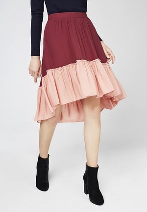 Vrouw draagt een tweekleurige bordeauxrode en roze gerimpelde high-low rok, gecombineerd met zwarte enkellaarsjes en een donker, lang mouwen top.