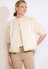 Crème-kleurige mouwloze vest, gemaakt van zachte faux bont met een gestructureerde afwerking, met een ronde kraag en vier knoopsluitingen.