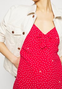 Czerwona sukienka w kropki z detalem w postaci kokardy na przodzie, z dekoltem w kształcie litery V. Modelka ma na sobie jasnokremową kurtkę dżinsową z mosiężnymi guzikami.