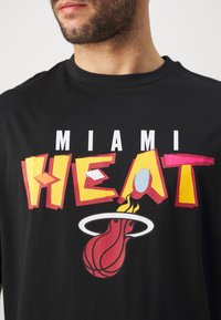 Camiseta de algodón negra con un gráfico audaz y colorido de "MIAMI HEAT", que incluye un diseño de llama y baloncesto. Mangas cortas y cuello redondo.