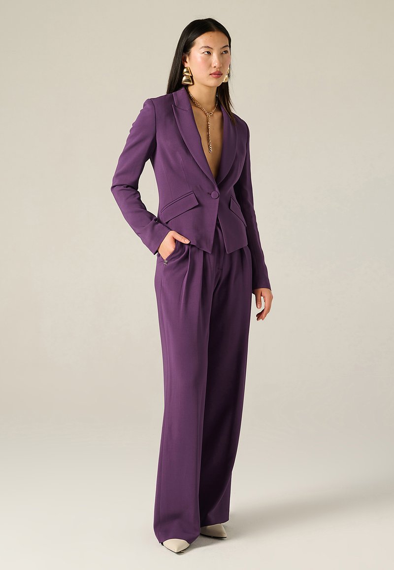 Blazer viola su misura con scollo a V profondo, due tasche frontali e chiusura con un bottone, abbinato a pantaloni a vita alta e gamba ampia.