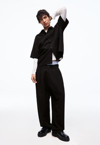 Ung mand iført lagdelte sorte og hvide toppe med brede sorte bukser og sorte sko, stående med den ene arm løftet bag hovedet.