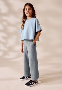 Camicetta azzurra chiara a maniche corte e cropped, pantaloni della tuta grigi a gamba larga, sneakers nere con righe bianche. Tessuto morbido e vestibilità comoda.