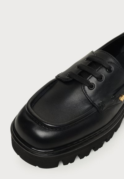 Chaussure en cuir noir à lacets avec semelle épaisse et rainurée et un design à bout carré, avec un petit élément décoratif doré sur le côté.