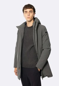 Giacca impermeabile grigia con cappuccio, dotata di zip frontale, due tasche laterali e un maglione grigio scuro a trama sotto.