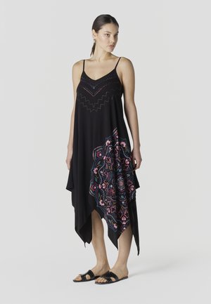 Mujer con un vestido negro sin mangas con un patrón de mandala colorido en un lado y sandalias negras tipo slide, de pie frente a un fondo liso.