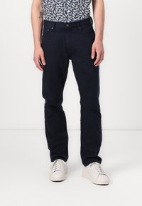 Pantaloni in cotone blu navy con taglio dritto, dotati di tasche laterali e chiusura con bottone metallico, indossati con sneakers bianche.