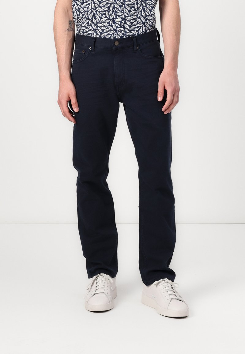 Pantaloni in cotone blu navy con taglio dritto, dotati di tasche laterali e chiusura con bottone metallico, indossati con sneakers bianche.