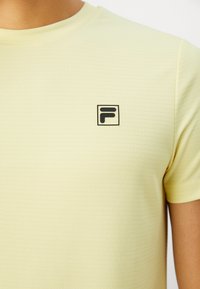 Nærbilde av en person som har på seg en lys gul teksturert T-skjorte med en liten svart og hvit "F" logo på venstre bryst.