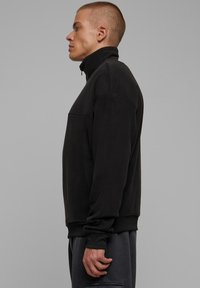 Svart fleece-pullover med høy krage, glidelås ved halsen, og ribbestrikkede mansjetter og kant. Glatt tekstur, enkelt design og avslappet passform.