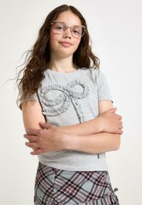 Grå kortærmet T-shirt med et tekstureret flæse-design i form af en sløjfe foran. Mønstret ternet nederdel ses nedenfor.