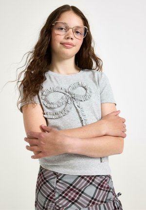 Grå kortärmad T-shirt med en texturerad volangdesign i form av en rosett framtill. Mönstrad rutmönstrad kjol synlig nedanför.