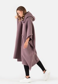 Przekroczony, z kapturem poncho z miękkiej mauve tkaniny. Wyposażony w kieszeń kangura, bezszwowe krawędzie i luźny krój. Noszony na czarnych legginsach i białych sneakersach.