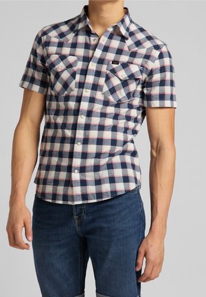 Mand iført en kortærmet, blå og hvid ternet skjorte med brystlommer og mørkeblå jeans mod en ensfarvet baggrund.