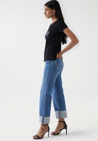 Schwarzes T-Shirt mit kurzen Ärmeln und blauem Grafiktext. Hellblaue, hochgeschnittene Jeans mit umgeschlagenen Säumen. Schwarze, sandalenartige High-Heels mit Riemchen.