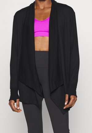 Cardigan nero aperto sul davanti indossato sopra un reggiseno sportivo rosa acceso e leggings neri a vita alta su una persona.