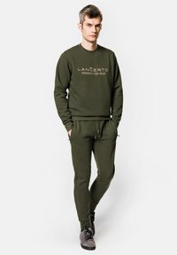 Oliwkowy sweatshirt i dresy wykonane z miękkiego materiału. Sweatshirt ma nadrukowane logo; dresy posiadają boczne kieszenie i sznurek w pasie.