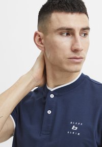 Tamsiai mėlynas polo marškinėliai su tekstūruota medžiaga, balta apykakle ir dviem mygtukais. Krūtinėje įspaustas baltas logotipas "BLEND DENIM".