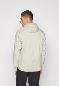 Lys beige hættejakke med et glat, vandafvisende stof, der har lange ærmer og en kant, der falder lidt under taljen.